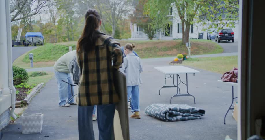 The video shows a group of four people setting up white folding tables on a driveway viewed from inside a garage or doorwaya woman in a plaid shirt and three other individuals are shown collaborating to unfold and place a large table with two other