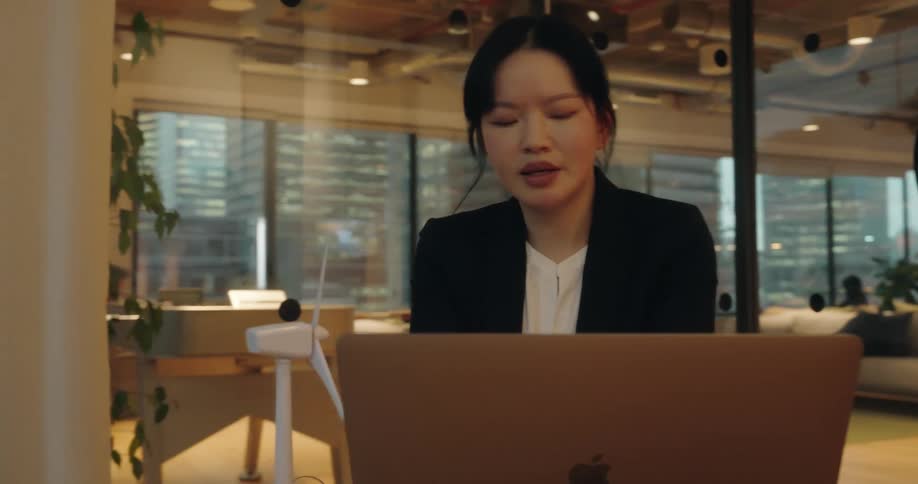 A woman wearing a blazer sits at a table in a glasswalled office speaking while looking at a laptop screen next to a small model wind turbine
