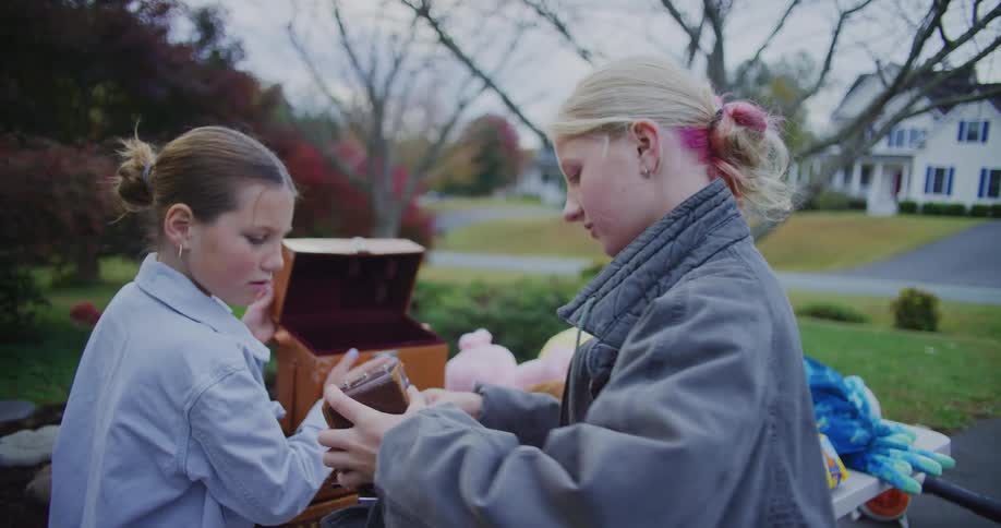 Two teenage girls open boxes and examine items with one showing an excited expression while holding a small vintage camera outdoors in a suburban driveway