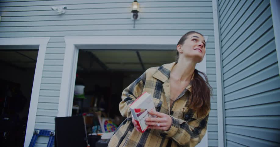 A young woman in a beige and black plaid jacket stands outside a garage holding a small book and smiling as she looks upward and interacts with people nearby