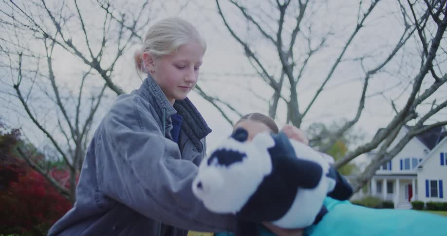Two young girls are outdoors possibly in a suburban yard helping to pack away large brightly colored inflatable items they handle stuffed animals including a black and white cow and a yellow duck pushing them into a blue bag