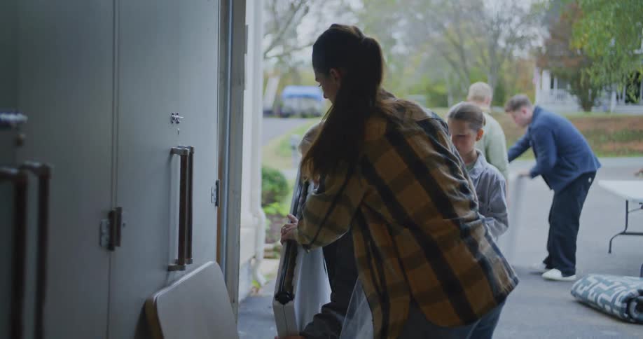 A young woman in a plaid shirt and jeans helps a group of people carry and set up folding tables and supplies in an outdoor setting for an event