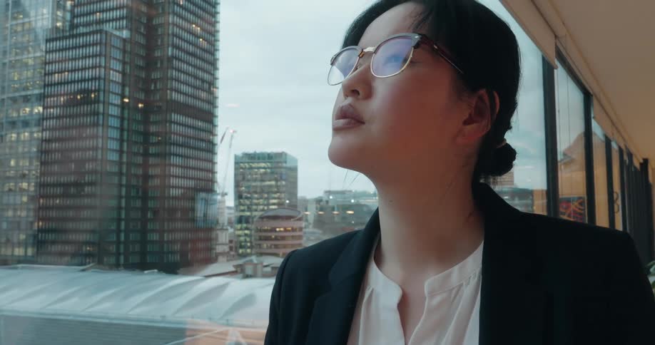 A serious woman wearing a black blazer and glasses holds and examines a small model of a wind turbine near a large office window overlooking a city skyline