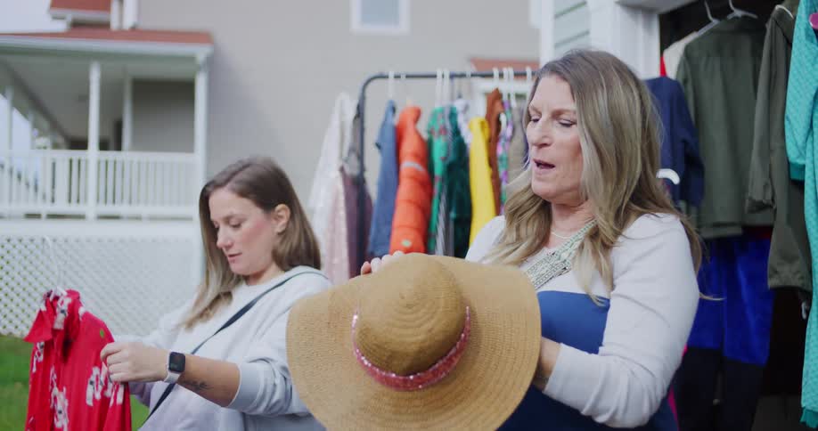 Two adult women are browsing through clothes at an outdoor rack with one woman holding a widebrimmed straw hat and talking animatedly while the other woman examines a red patterned dress