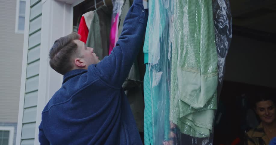 A young man in a dark blue jacket is reaching up to a temporary rack of clothes which is hanging under an open garage door or awning he pulls out a sparkly bright green garment that is protected inside a clear plastic drycleaning bag