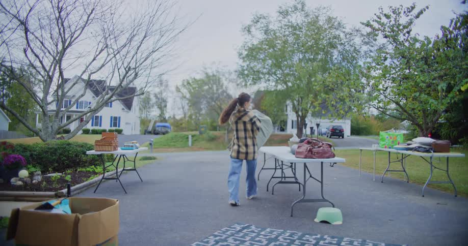 A woman wearing a plaid shirt and jeans sets up for an outdoor yard sale in a suburban driveway placing items onto folding tables and carrying a large bag