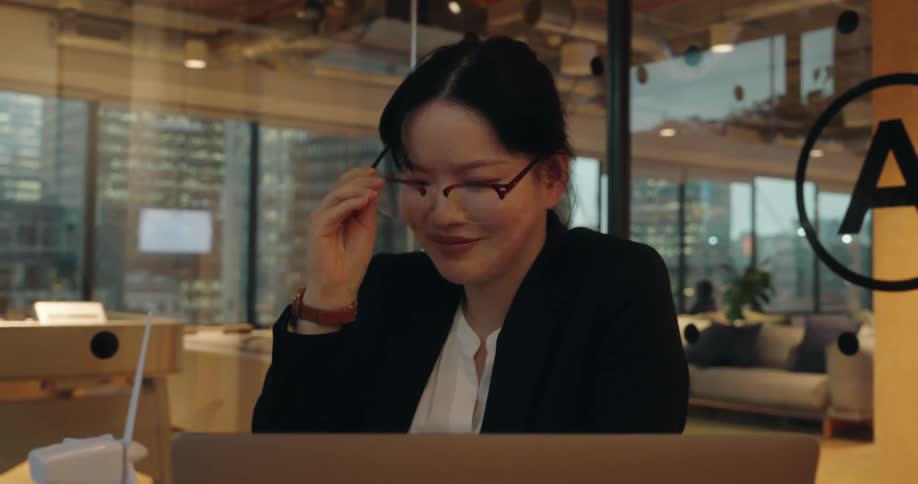 A professional woman in a suit and glasses participates in a video call on her laptop smiling and talking in a modern office with a city view at dusk