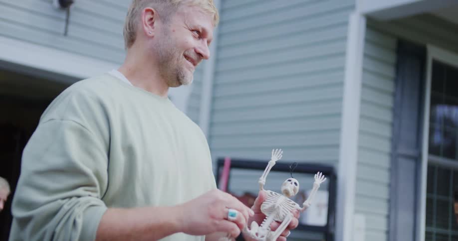 A close view of a smiling man in a light green sweatshirt holding a small plastic skeleton decoration outside a house with a woman visible in the blurred background