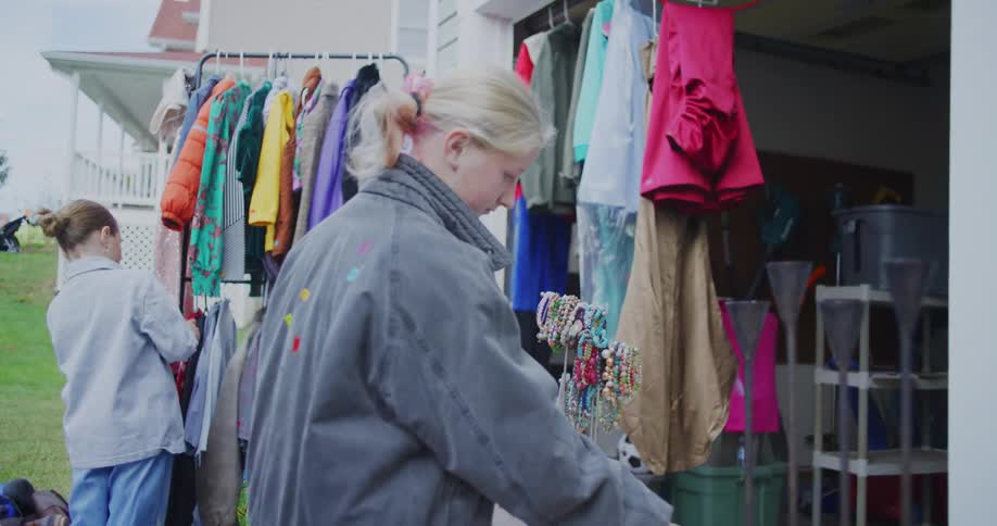 Two women browse items at a suburban garage sale where racks of colorful clothes and a stand of jewelry are set up outside a house