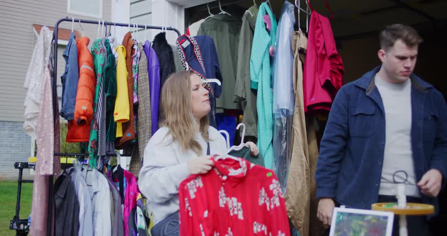 This short video clip features two people a man and a woman standing outside a garage that is set up for a yard sale specifically for clothes the woman is holding up a bright red patterned shirt and a dark green jacket on hangers possibly comparing t