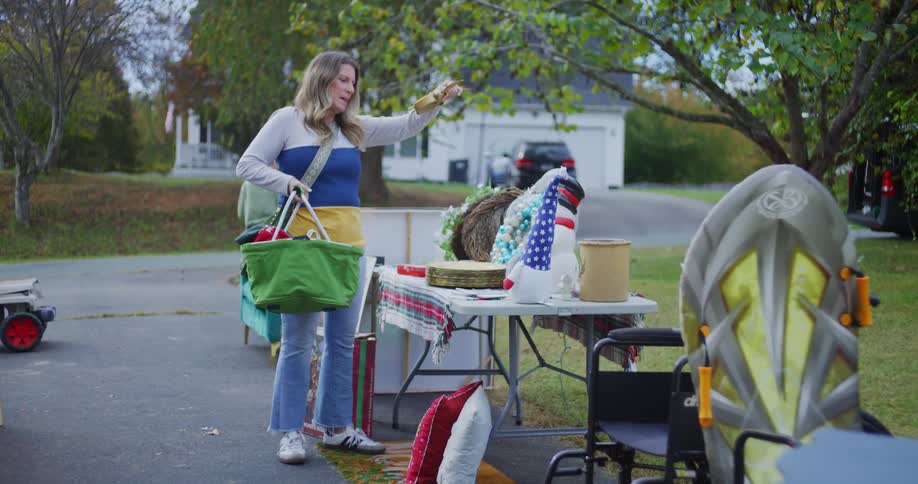 A middleaged woman holds a basket while browsing and picking up items at a garage sale under the bright midday sun