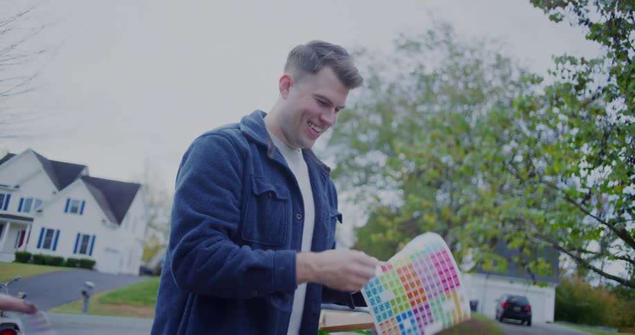 Two men are smiling and discussing a color palette card over a table full of garage sale items including a stuffed sloth and a woven wreath outside a suburban home