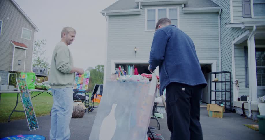 Two men are standing on a driveway in front of a house looking at a painting that is sitting among many items on the driveway