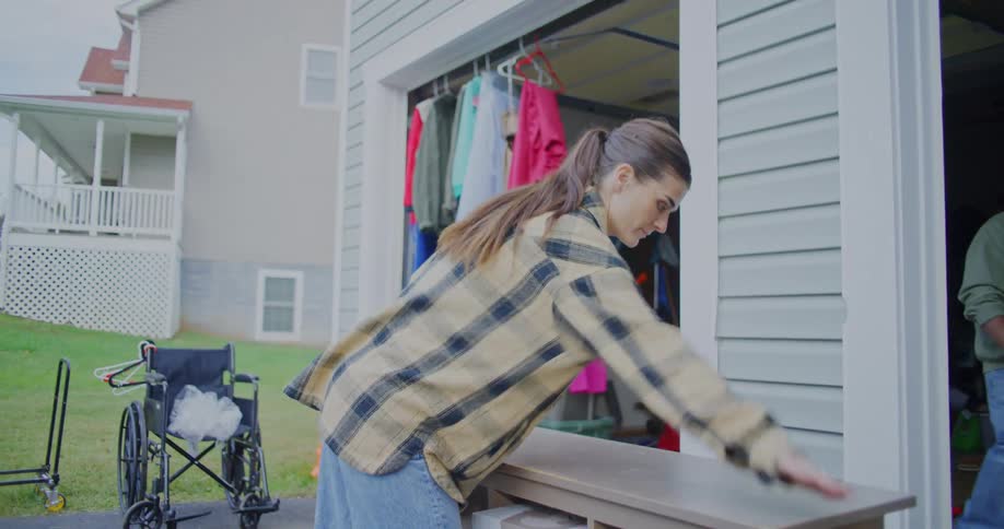 A young woman cleans a shelf outside a garage at midday preparing for a garage sale clothes hang neatly on the garage door while a man walks inside adding to the busy setup scene