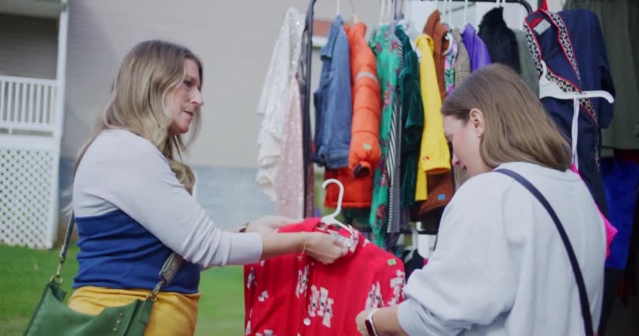 Two women examine a red floral dress at a yard sale clothing rack