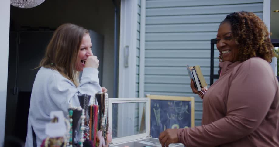 Two women are in a yard near a house laughing near jewelry and other items are on a table that looks like a garage sale