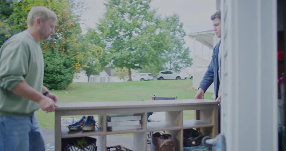 Two men carry a wooden shelving unit outside during a yard sale