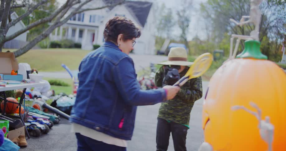 A young person wearing a straw hat and camouflage hoodie is seen holding a video camera and looking up at an older woman the woman wearing a denim jacket turns around and walks toward the camera personin the foreground to the right is a large inflat