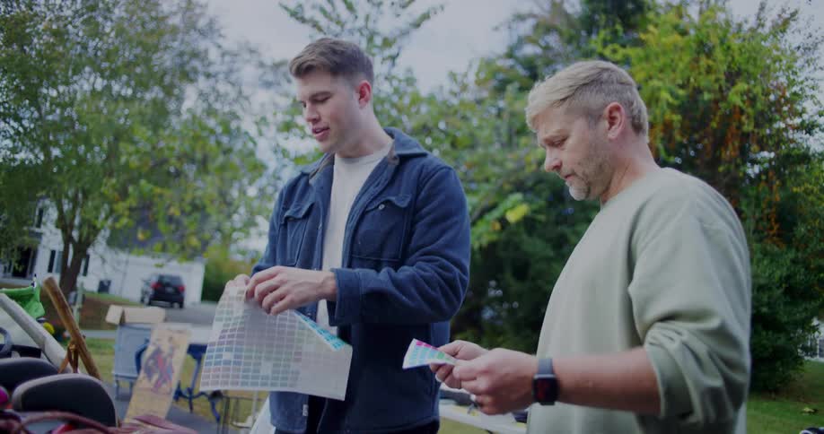 Two men organize items including a color swatch grid at an outdoor yard sale