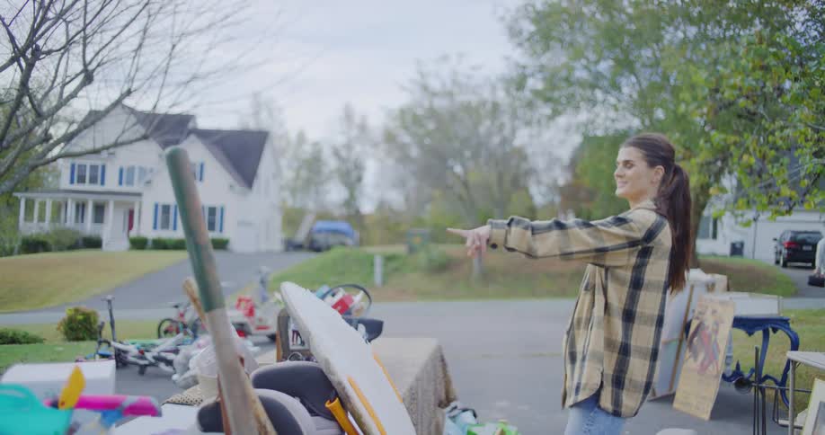 A man and a woman are organizing items at what appears to be a garage sale with a wide assortment of goods displayed on a drivewaythe woman wearing a plaid shirt points offcamera then helps the man remove a yellowishorange door mat with a festiv