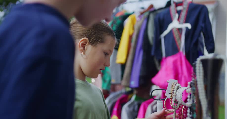 A closeup view focuses on two young girls closely examining a white display stand filled with various beaded and chained jewelry set against a colorful cluttered rack of clothing in the background