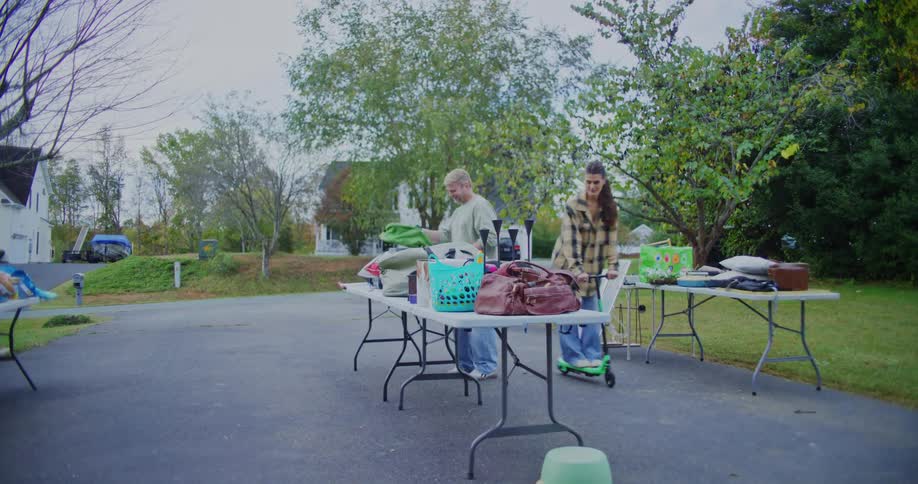 A young woman playfully rides a green electric scooter through a driveway yard sale weaving past folding tables laden with goods while the farther fold clothes before walking out of the scene