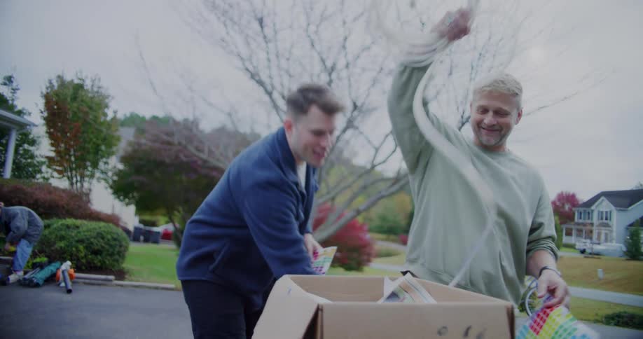 Two men smile and chat while sorting through white tubing and household items in a cardboard box at a yard sale