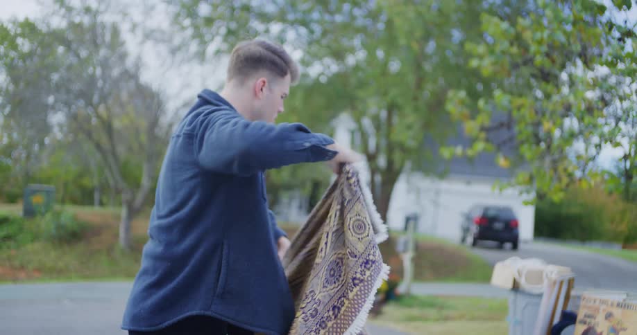 A man wearing a blue jacket unloads items from a large bag onto a folding table and then unfolds a patterned rug outdoors for a yard sale