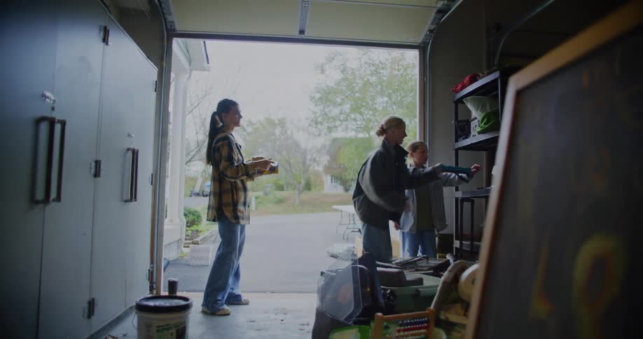 A group of people stand in the open doorway of a messy garage holding various items as they prepare for a yard sale on an overcast day outside a suburban home