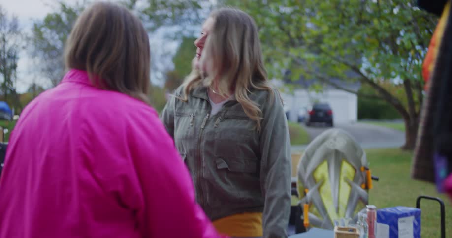 A woman trying a green jacket speaking animatedly to another woman in a bright pink jacket gesturing with her hands at an outdoor yard sale in a suburban setting