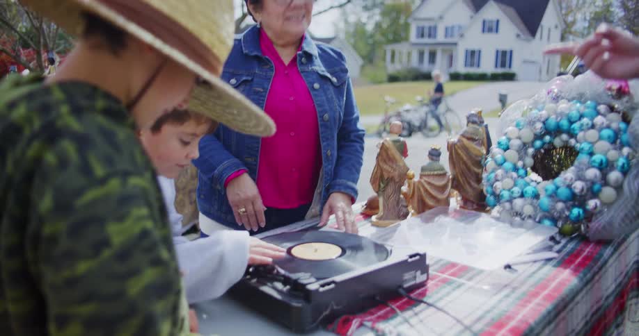An older woman in a denim jacket and a young boy are interacting with a portable record player on a plaidcovered table possibly listening to a record together at a community event or yard sale christmas decorations including three wise men statues and