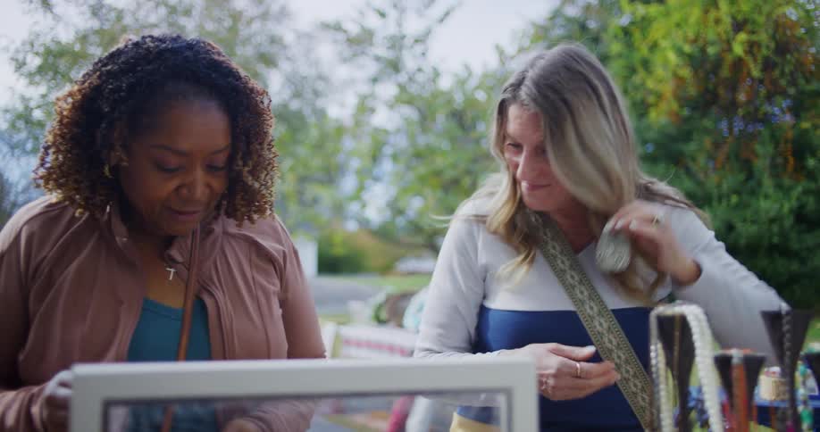 Two women browse items at an outdoor flea market or garage sale with one trying on a dangling earring and smiling in a casual outdoor setting