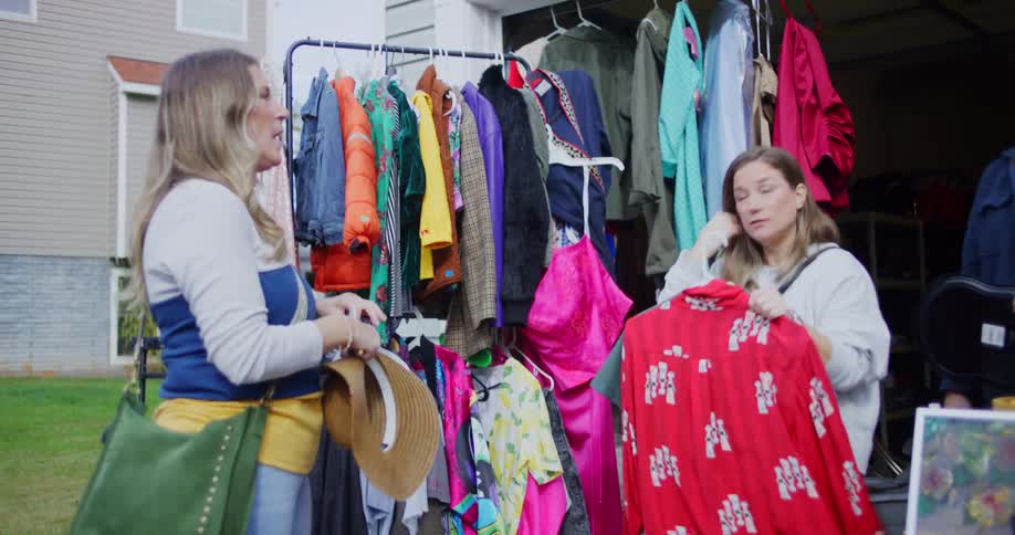 Two women browse clothing and a hat on an outdoor rack at a garage sale