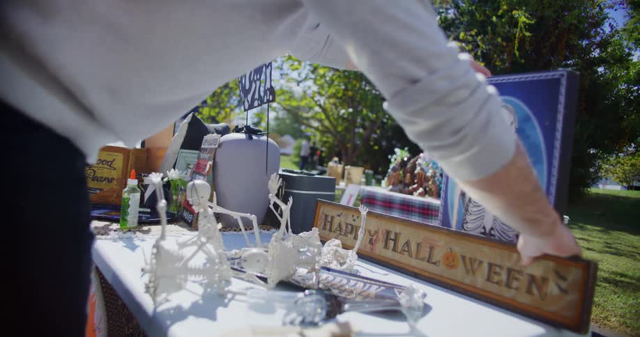 A close view shows a person arranging various halloween decorations including small skeletons and a framed skeleton image on a table at an outdoor yard sale