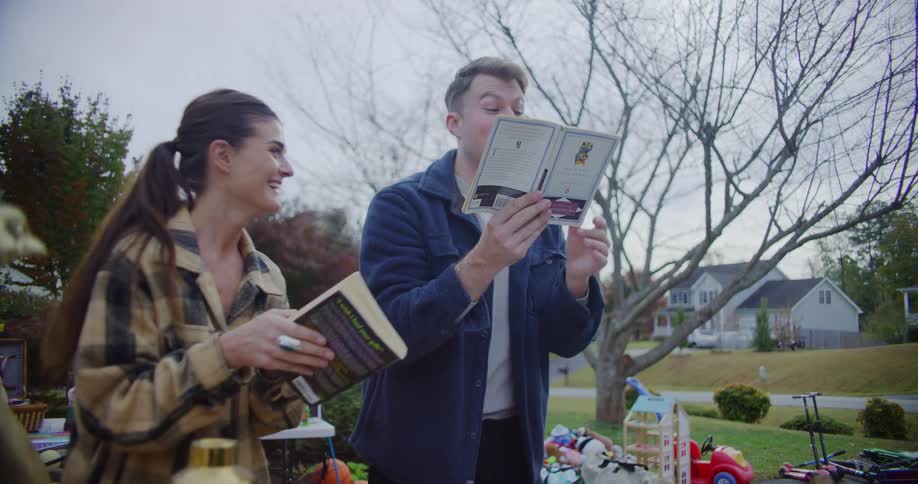 A couple laughs together while looking through old books they found at an outdoor yard sale in a suburban neighborhood on an overcast day
