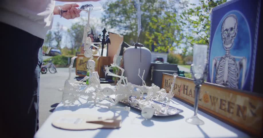 A person is setting up a table of small halloweenthemed decorations primarily miniature white plastic skeletons outdoors on a sunny day possibly at a yard sale or market they are carefully arranging the items which also include a framed skeleton por