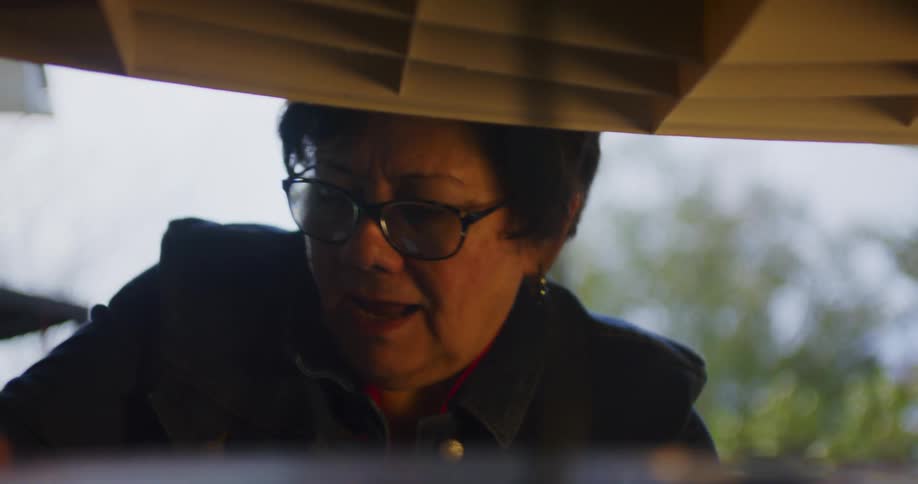 A close low angle view shows an older woman in glasses and a dark jacket smiling while interacting with a young boy over an object seen from underneath a wooden surface