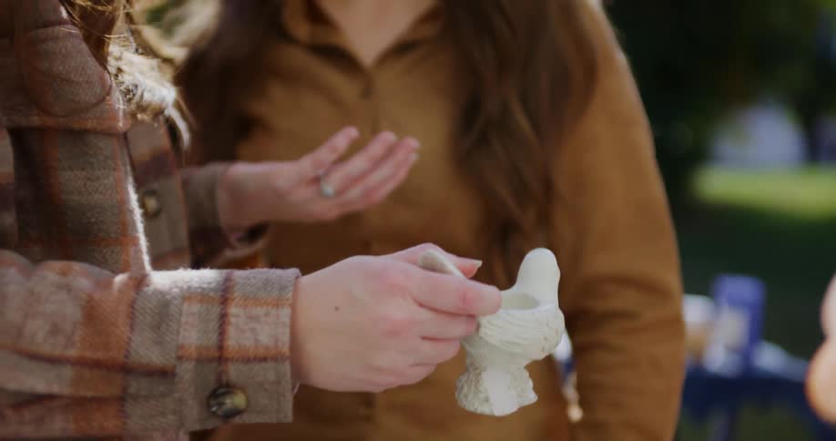 Two friends stand outdoors in the sunlight carefully dropping small beads into a hollow ceramic bird before looking up to smile at one another