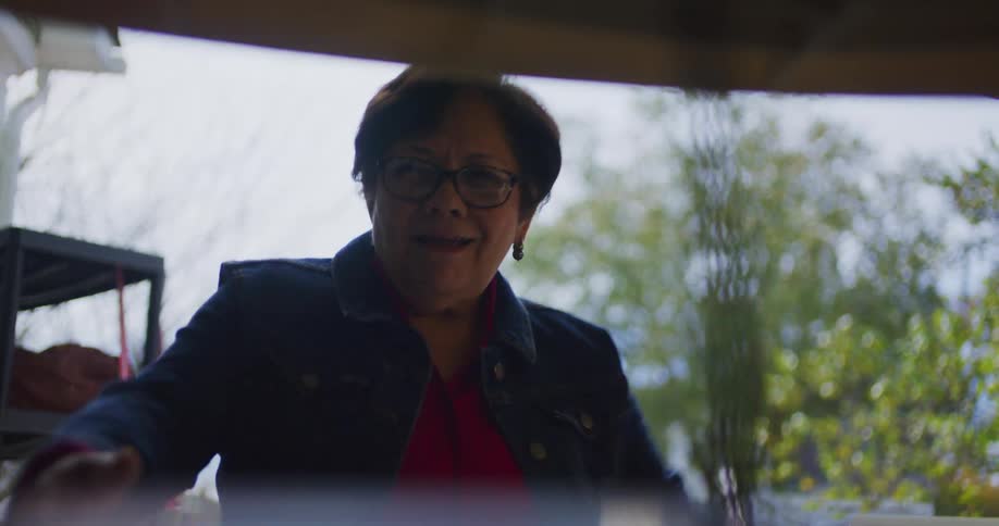 An older woman with glasses is outside looking surprised at first then smiling as she approaches the camerashe is bending down looking through or under an object in the foreground perhaps examining items at a yard sale or similar outdoor market