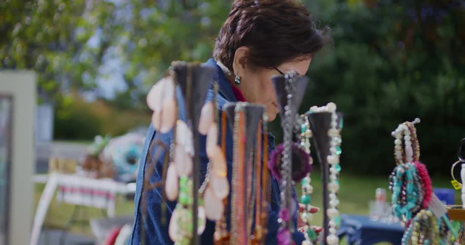 An older woman wearing glasses a pink shirt and a denim jacket intently examines jewelry displayed on racks at an outdoor market on a sunny day