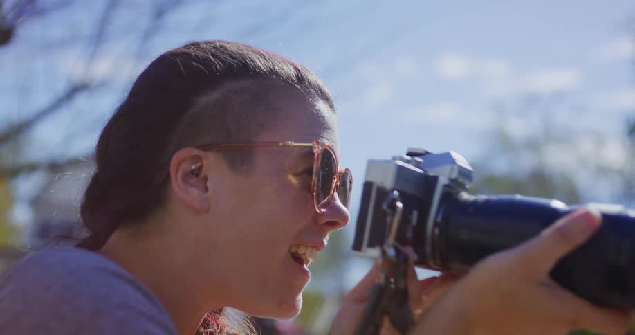 Bathed in bright autumn sunlight a photographer with a distinctive sideshave hairstyle twists the lens cap off her vintage camera laughing joyfully as she snaps a photo near a bright orange pumpkin