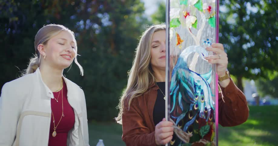 A younger woman with blonde hair wearing a white jacket over a maroon top is standing on the left and smiling as she looks at the piece of artwork the other woman is holdingan older woman with long blonde hair wearing a brown jacket is holding up a