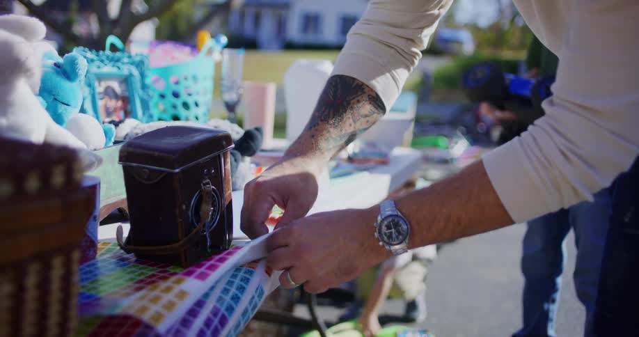 Young adult man putting price tags on objects over a table on a yard sale next to two kids outdoors during day time