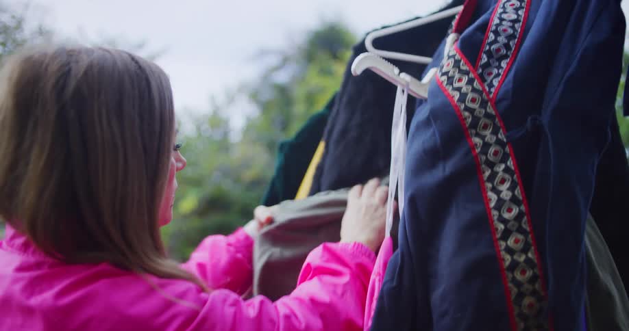 A young woman picks a dress from a hanger filled with clothes and hands it to another woman while shopping outdoors at a garage sale