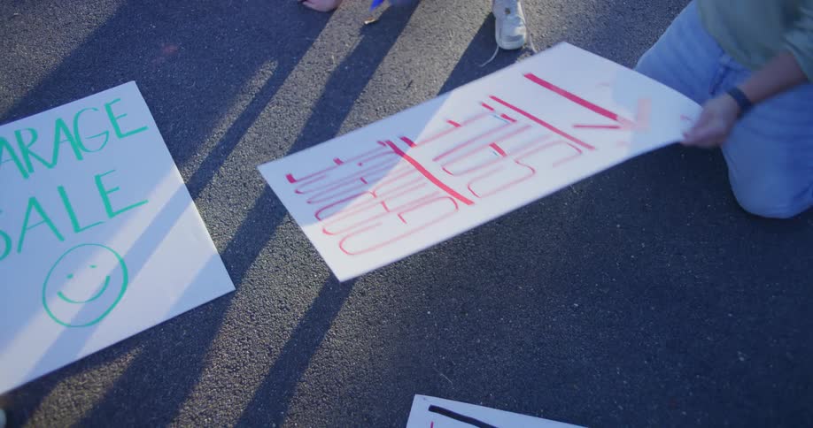 Two people kneel on asphalt making handmade garage sale signs for a neighborhood event