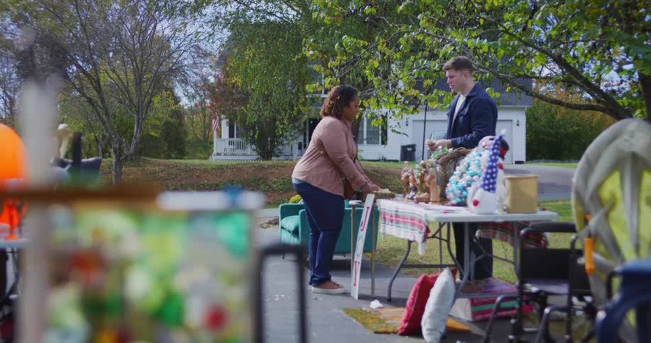 This video shows a woman and a young man talking at an outdoor yard or garage sale on a sunny day the woman is holding a small brown box seemingly having just purchased an item while the man gestures toward a table filled with various festive trinkets