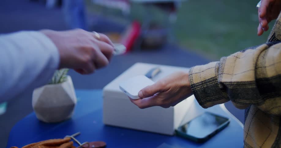 A close view shows a transaction taking place as a person uses a mobile phone to tap and pay with a digital wallet to another person holding a small white payment device over a cash box