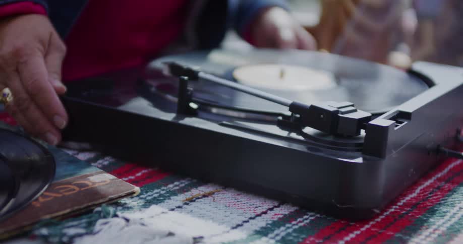 A persons hands are shown operating a black vinyl record player they gently lift the needle from the spinning record then lower the arm again likely to stop or restart the musicthe record player is sitting on a patterned surface possibly a blanke