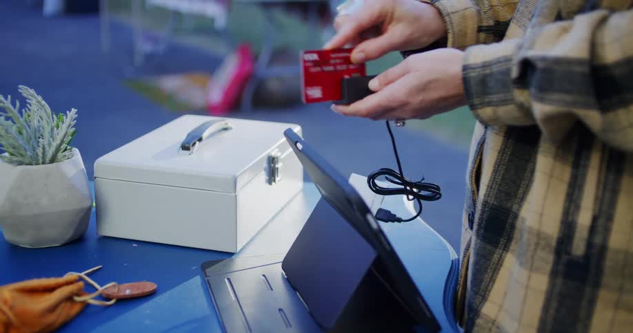 A woman stands in front of a desk outdoors at midday collecting a credit card and sliding it through a payment processor to complete the transaction