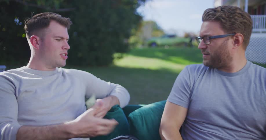 Two men are sitting outside on a couch with green cushions the man on the right wearing glasses and a gray tshirt extends a wad of cash to the man on the left who is wearing a light gray longsleeved shirt